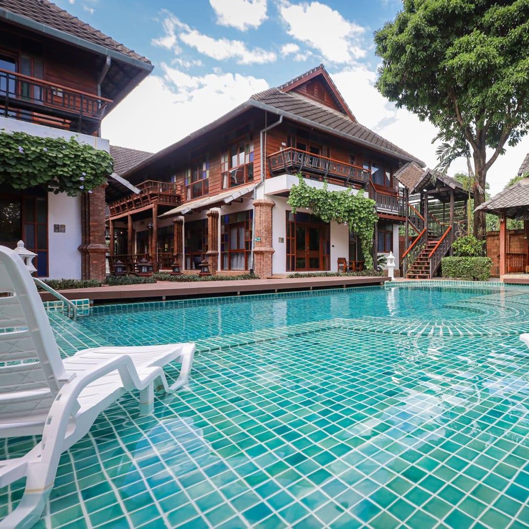 The pool, white loungers, a garden sala and a teak building under a blue sky at Jintara Rehab in Chiang Mai