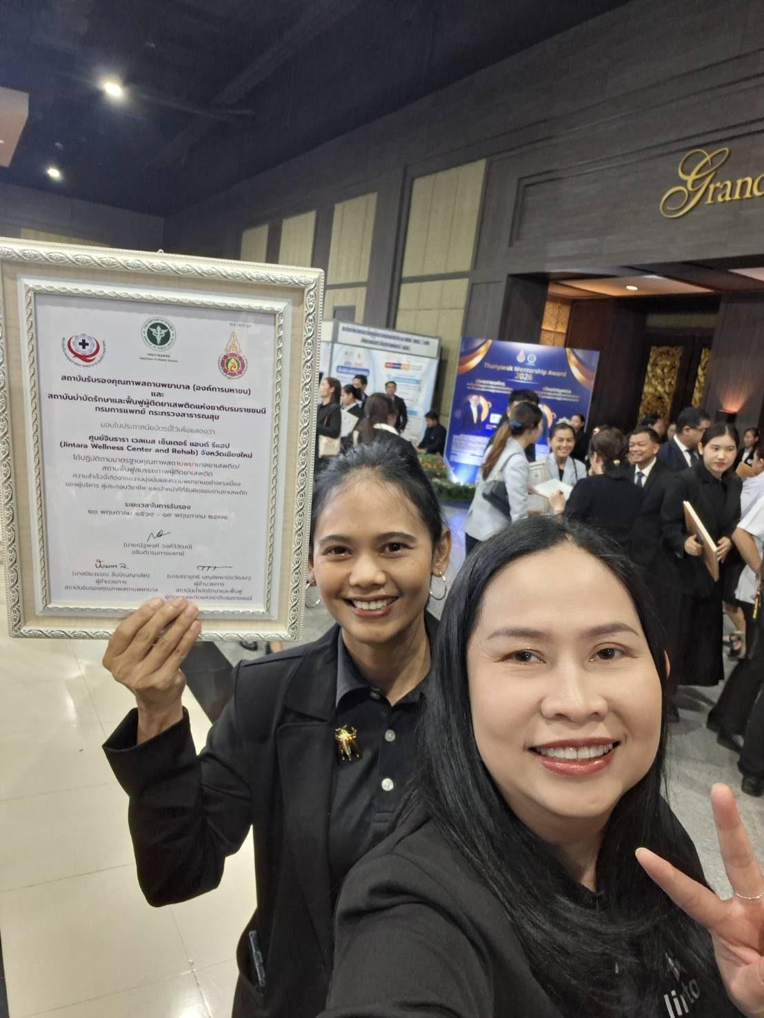 Two Jintara Rehab staff smiling with the framed national accreditation certificate at the conference venue in Bangkok