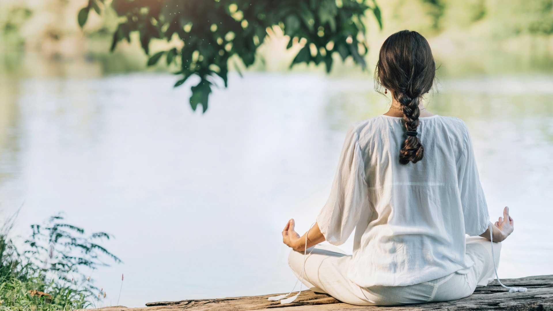 Woman meditating cross-legged by a calm river surrounded by greenery at Jintara Rehab